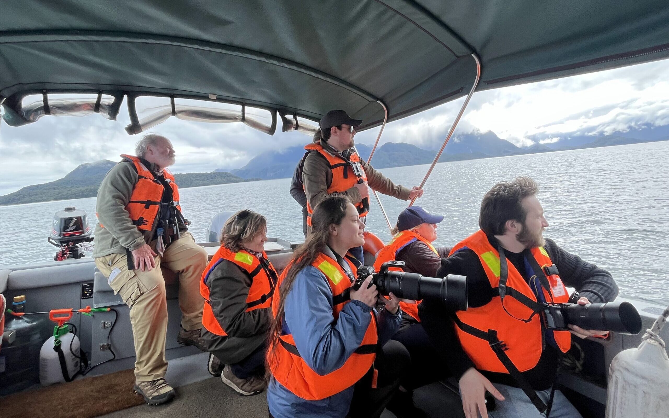 IMG_3844 A group of nature guardians, wearing orange lifejackets, look over the edge of a boat into the water. The two guardians in the front of the group each hold a large camera.