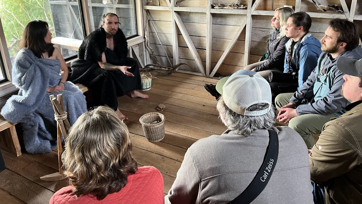 A group of people is seated in a rustic wooden room, attentively listening to two individuals who are speaking. The the speakers are wearing furs. The atmosphere appears to be informal and engaged.