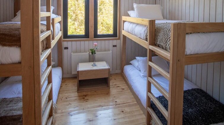 A bunk room with two sets of bunkbeds, one on each side of a window. A small table stands below the window.